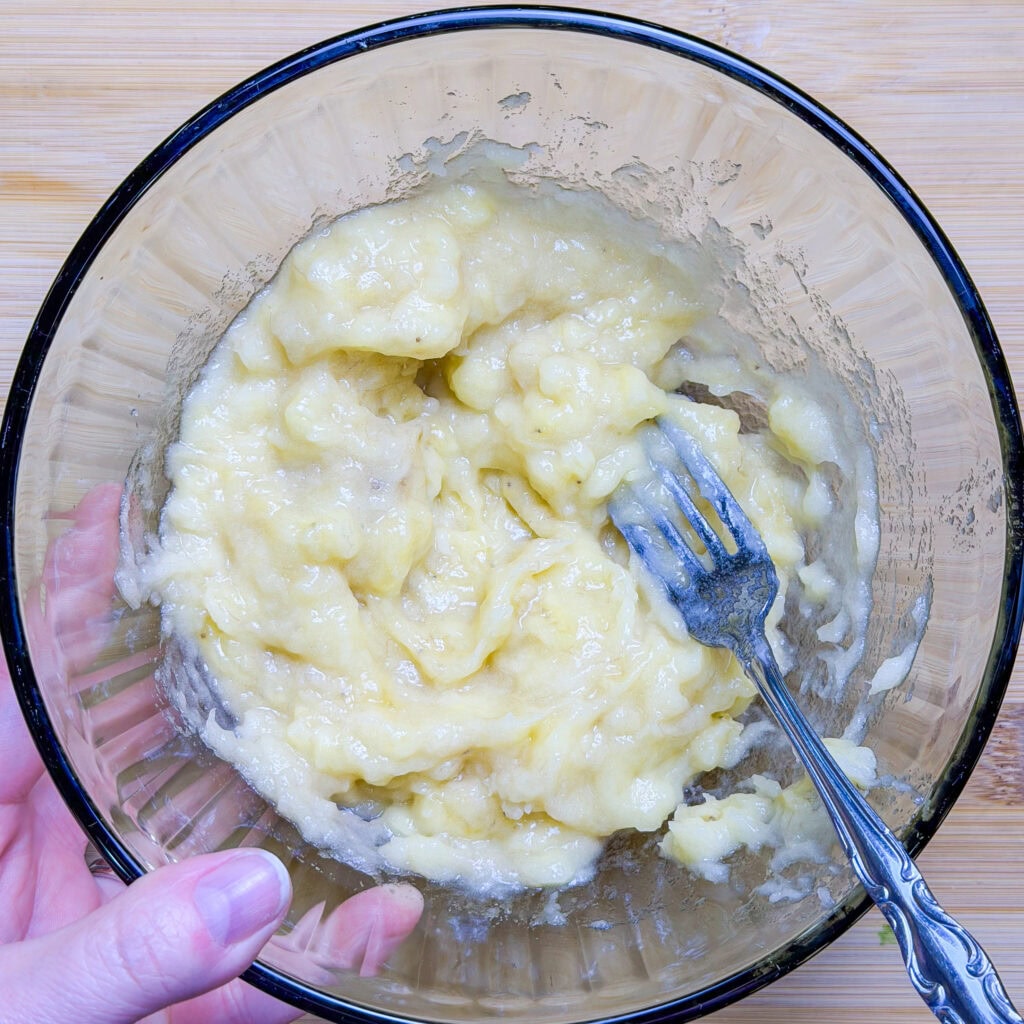 A hand holds a glass bowl with mashed bananas inside. A fork, used for mashing, rests in the mixture. The bowl sits on a wooden surface.
