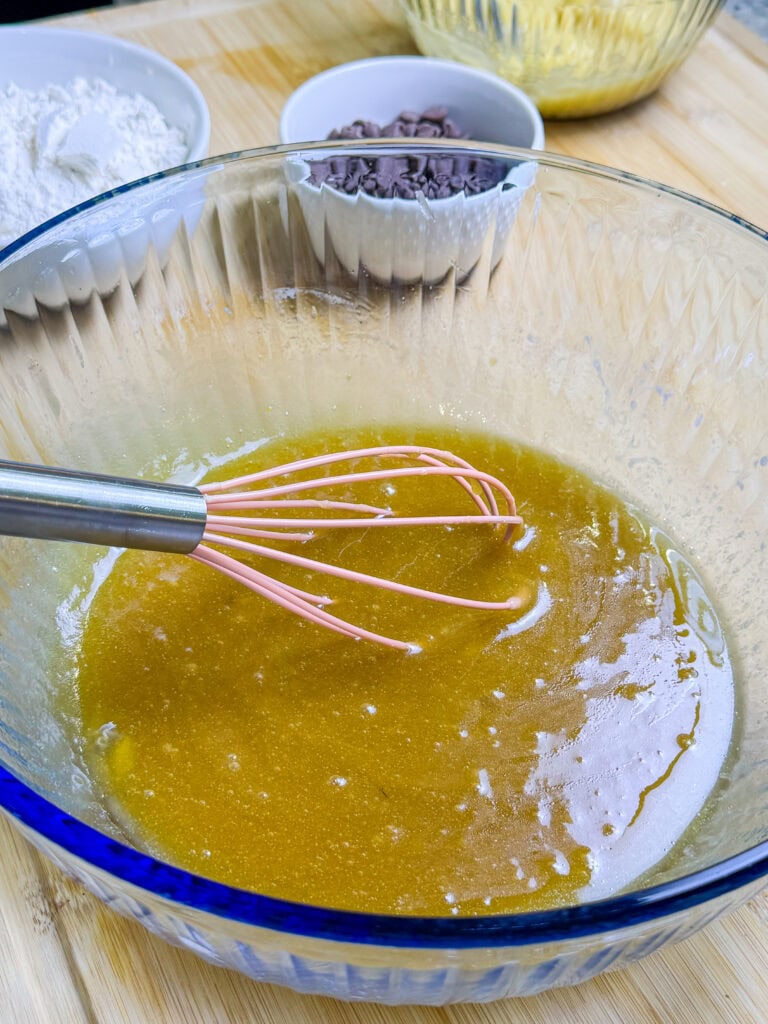 A glass bowl with a metal whisk mixing wet ingredients, likely for baking. In the background are bowls containing flour and chocolate chips on a wooden surface.