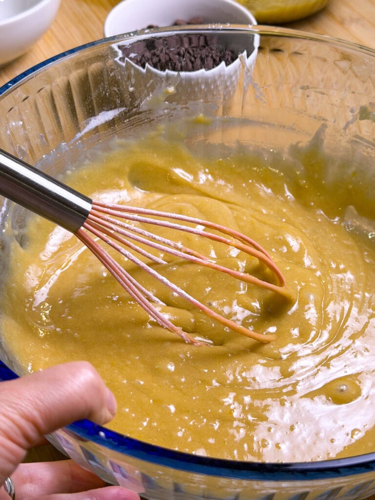 A hand holds a whisk mixing yellow batter in a large glass bowl, with chocolate chips and other ingredients in small bowls in the background on a wooden surface.