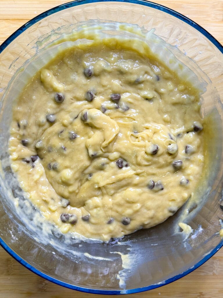 A glass mixing bowl filled with thick banana bread batter mixed with chocolate chips, sitting on a wooden surface. The batter appears creamy with visible chunks and chocolate chips throughout.