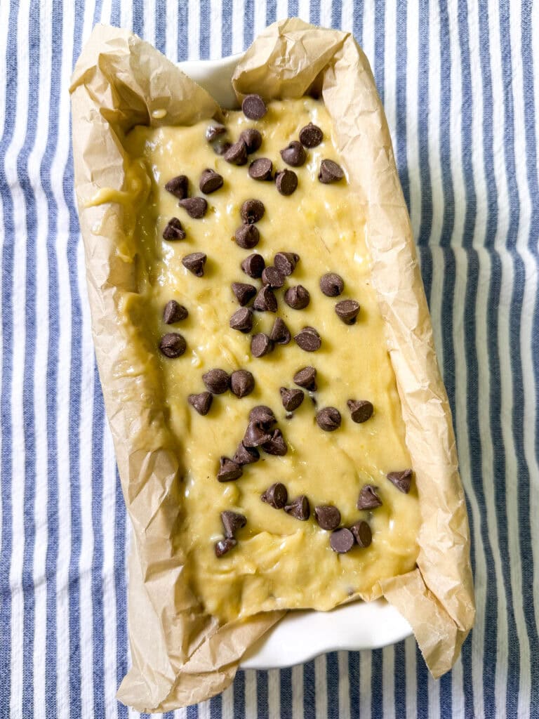 A loaf pan lined with parchment paper is filled with banana bread batter, topped with chocolate chips, and placed on a blue and white striped cloth.