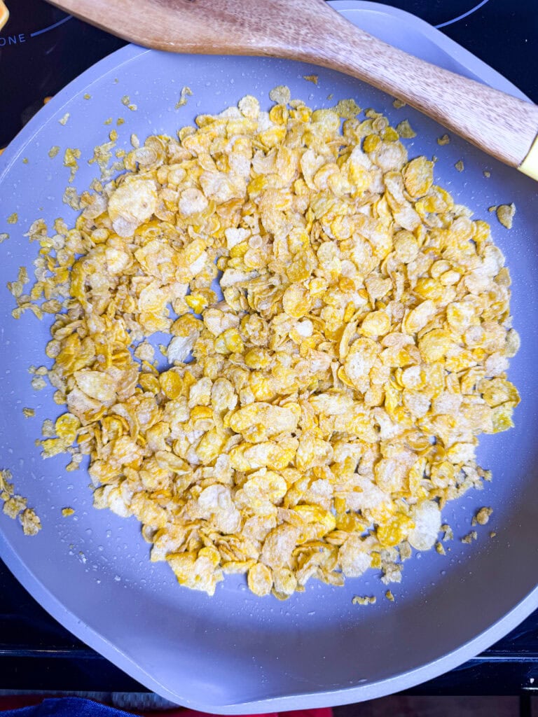 A frying pan filled with cornflakes, some broken into small pieces, with a wooden spoon resting on the edge of the pan.