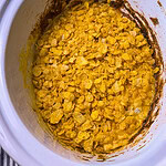 A slow cooker filled with a baked dish topped with golden cornflakes. The edges show some browned residue, suggesting the dish has been cooked or baked. The interior is creamy and topped with crunchy cereal.