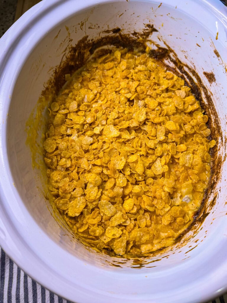 A slow cooker filled with a baked dish topped with golden cornflakes. The edges show some browned residue, suggesting the dish has been cooked or baked. The interior is creamy and topped with crunchy cereal.