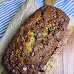A freshly baked loaf of banana bread topped with chocolate chips sits on parchment paper on a wooden cutting board, with a striped cloth partially visible underneath.