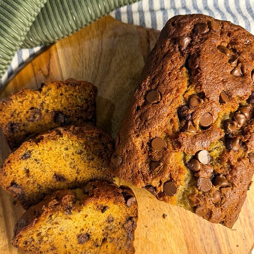 A loaf of chocolate chip banana bread sits on a wooden board, with three slices cut. The bread looks moist with chocolate chips sprinkled on top and throughout. A green and white striped cloth lies nearby.