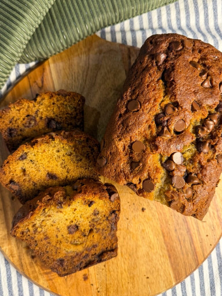 A loaf of chocolate chip banana bread sits on a wooden board, with three slices cut. The bread looks moist with chocolate chips sprinkled on top and throughout. A green and white striped cloth lies nearby.