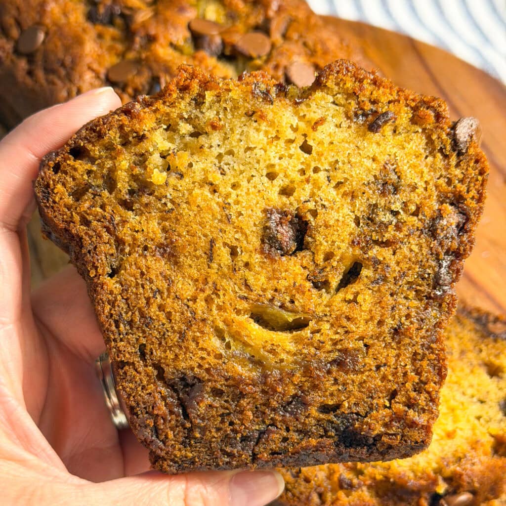 A hand holding a thick slice of moist banana bread with visible chocolate chips, showing a golden brown crumb and texture. More slices and a loaf are blurred in the background.