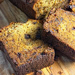 A close-up of a sliced loaf of chocolate chip banana bread on a wooden cutting board, showing the moist texture and scattered chocolate chips throughout the bread.