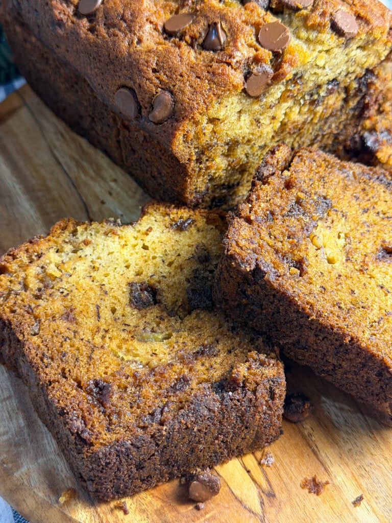 A close-up of a sliced loaf of chocolate chip banana bread on a wooden cutting board, showing the moist texture and scattered chocolate chips throughout the bread.