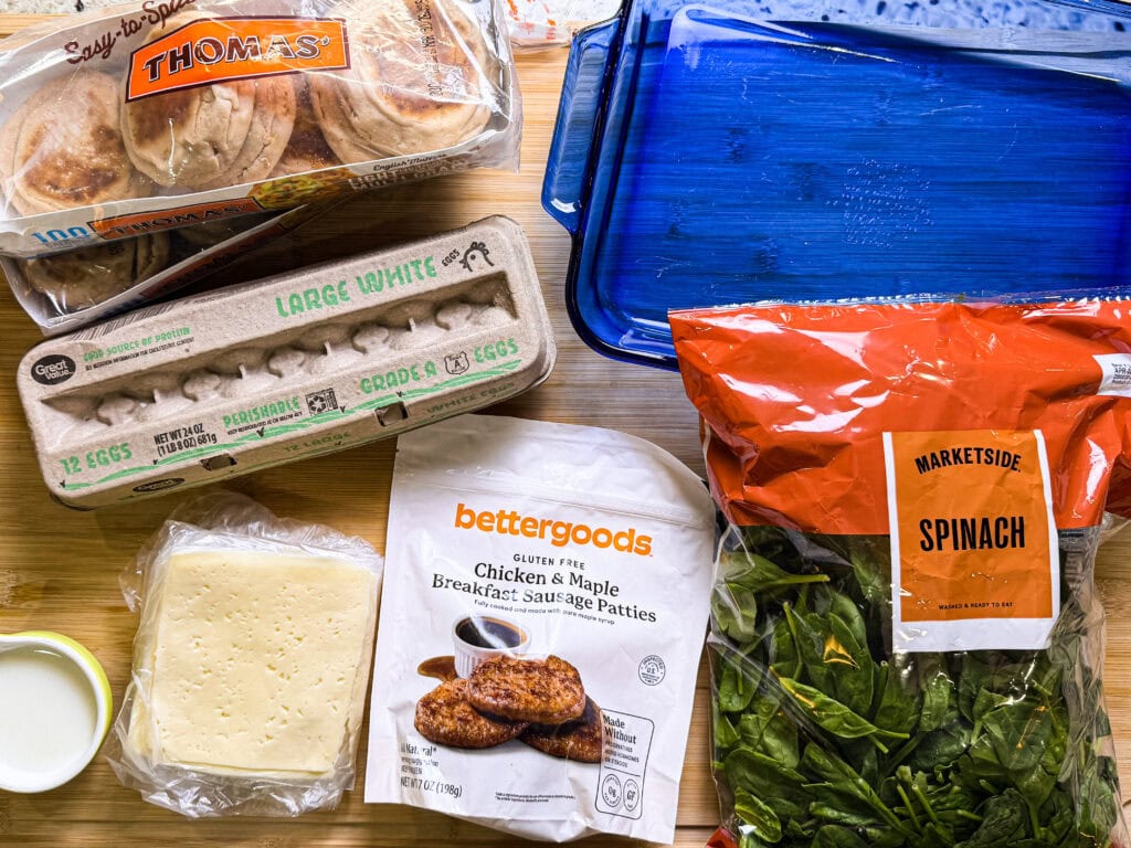 A variety of breakfast ingredients on a table, including eggs, sliced cheese, chicken & maple breakfast sausage patties, spinach, milk in a small cup, English muffins, and a blue baking dish.
