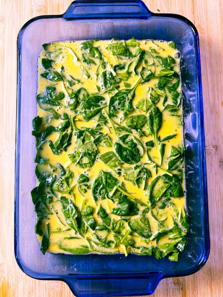 A rectangular blue glass baking dish filled with a mixture of raw spinach leaves and beaten eggs sits on a light wooden surface, ready to be cooked.