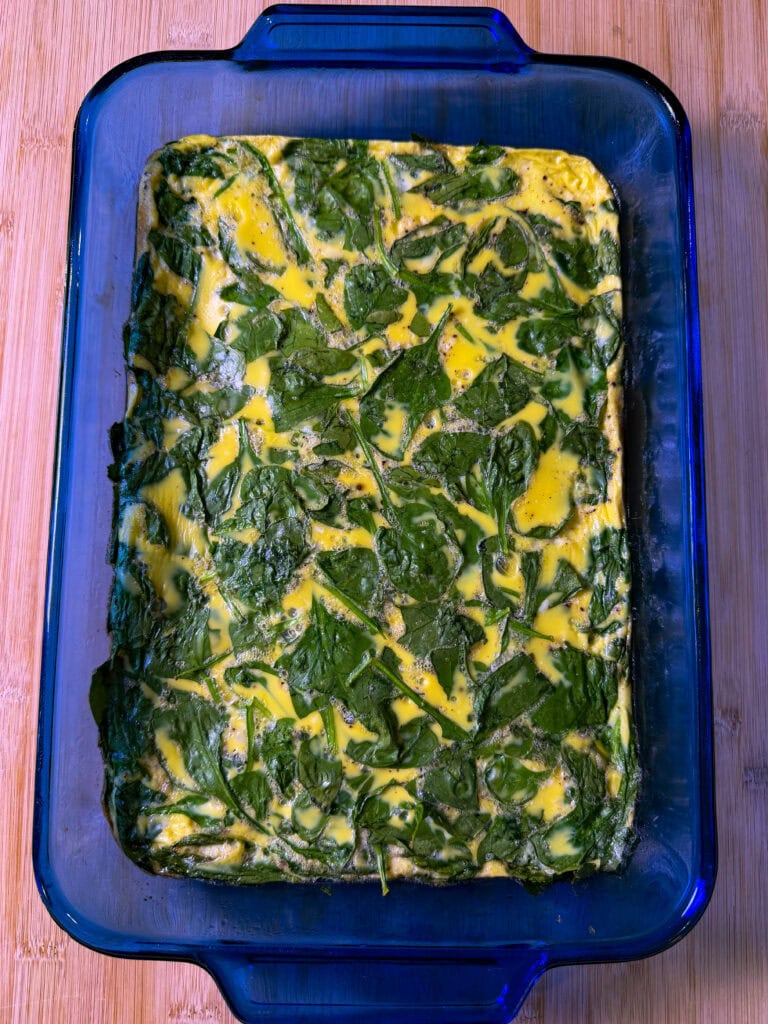 A blue glass baking dish contains a baked egg and spinach casserole, with visible green spinach leaves mixed into the yellow egg base. The dish rests on a wooden surface.