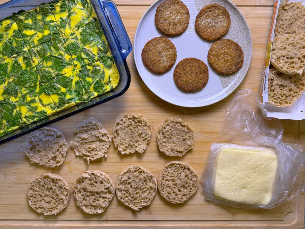 A top-down view of breakfast sandwich ingredients: a spinach and egg bake in a glass dish, sausage patties on a plate, sliced English muffins, and a stack of cheese slices on a wooden surface.