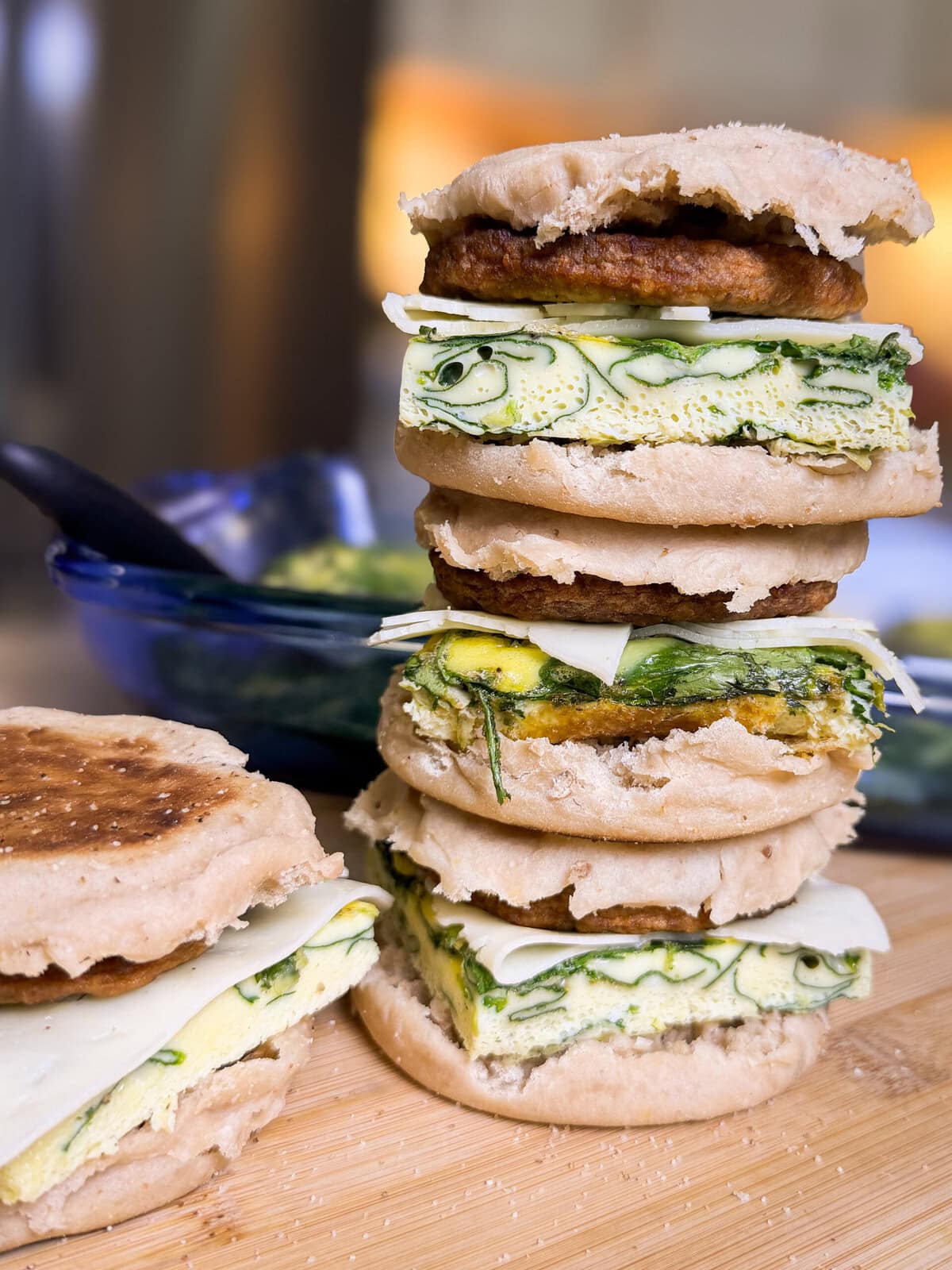 Three breakfast sandwiches stacked on a wooden board, each with English muffins, egg, spinach, cheese, and sausage. Another sandwich and a blue dish are blurred in the background.