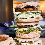 A close-up of three stacked breakfast sandwiches with English muffins, egg patty with green herbs, cheese, and sausage, on a wooden cutting board in a kitchen setting.