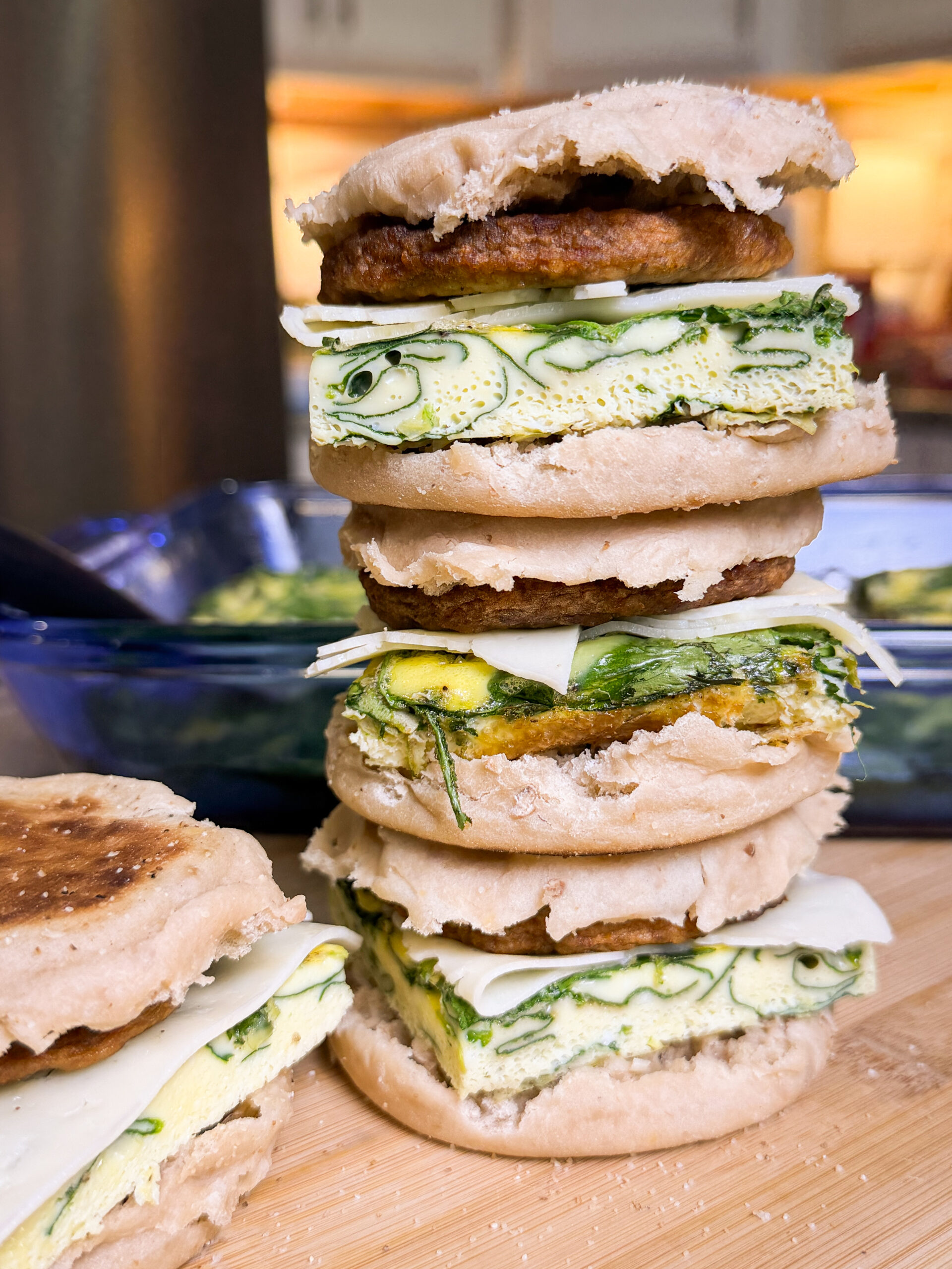 A close-up of three stacked breakfast sandwiches with English muffins, egg patty with green herbs, cheese, and sausage, on a wooden cutting board in a kitchen setting.