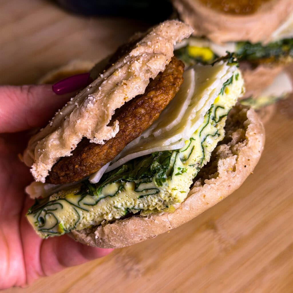 A hand holding a breakfast sandwich with an English muffin, sausage patty, shredded cheese, and a thick slice of spinach-filled egg on a wooden surface.