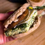 A hand holding a breakfast sandwich with an English muffin, sausage patty, shredded cheese, and a thick slice of spinach-filled egg on a wooden surface.