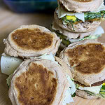 A close-up of several breakfast sandwiches with eggs and cheese on toasted English muffins, stacked on a wooden surface.