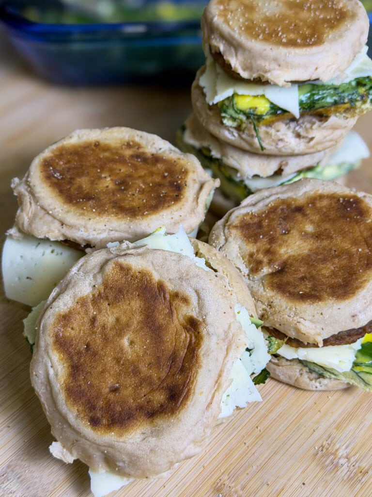A close-up of several breakfast sandwiches with eggs and cheese on toasted English muffins, stacked on a wooden surface.