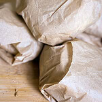 Three sandwiches or food items wrapped in brown paper are stacked on a wooden surface. The paper looks crumpled and the items are arranged closely together. No labels or markings are visible.