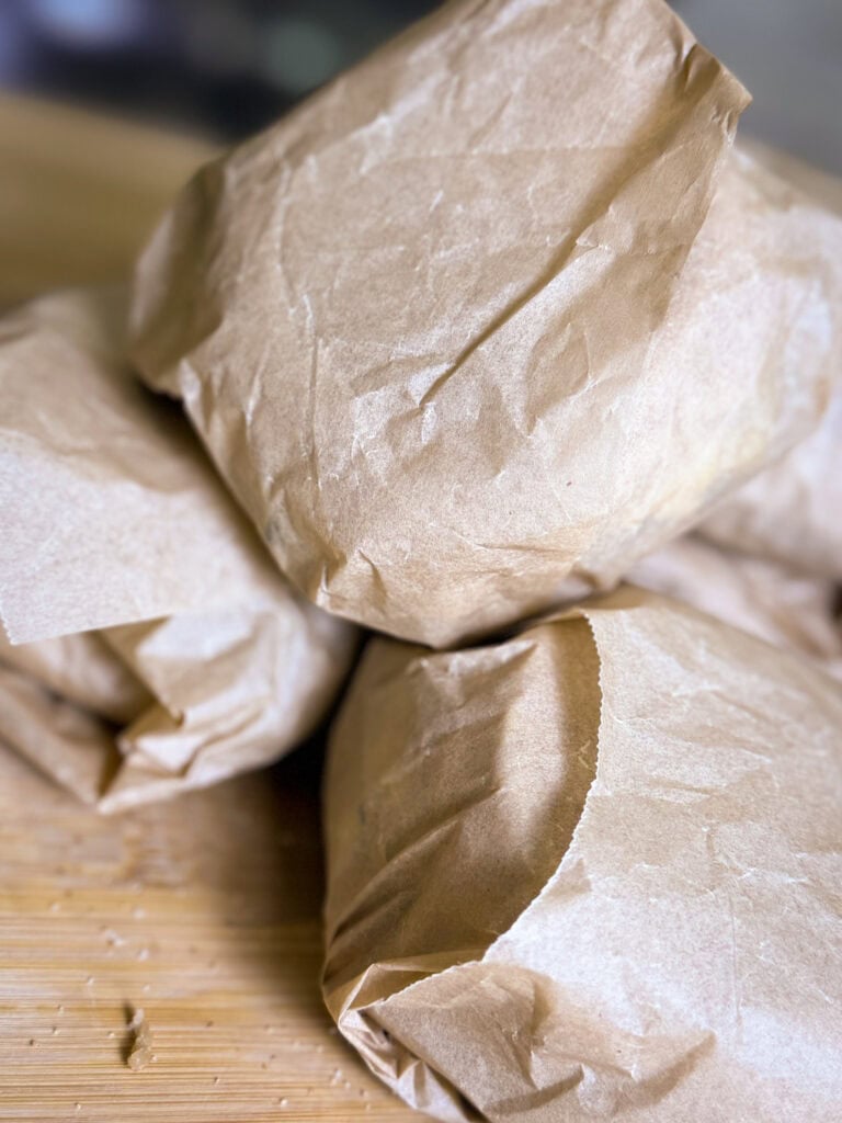 Three sandwiches or food items wrapped in brown paper are stacked on a wooden surface. The paper looks crumpled and the items are arranged closely together. No labels or markings are visible.