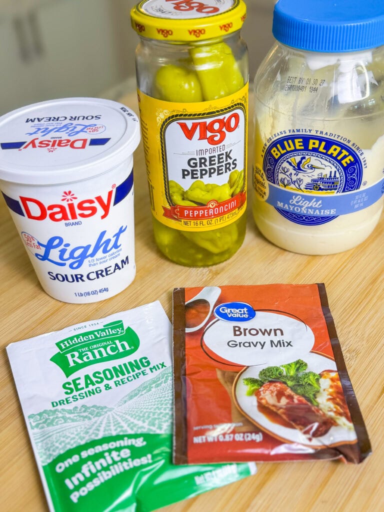 Five ingredients on a wooden surface: Daisy Light sour cream, Vigo Greek pepperoncini, Blue Plate Light mayonnaise, Hidden Valley Ranch seasoning packet, and a packet of brown gravy mix.