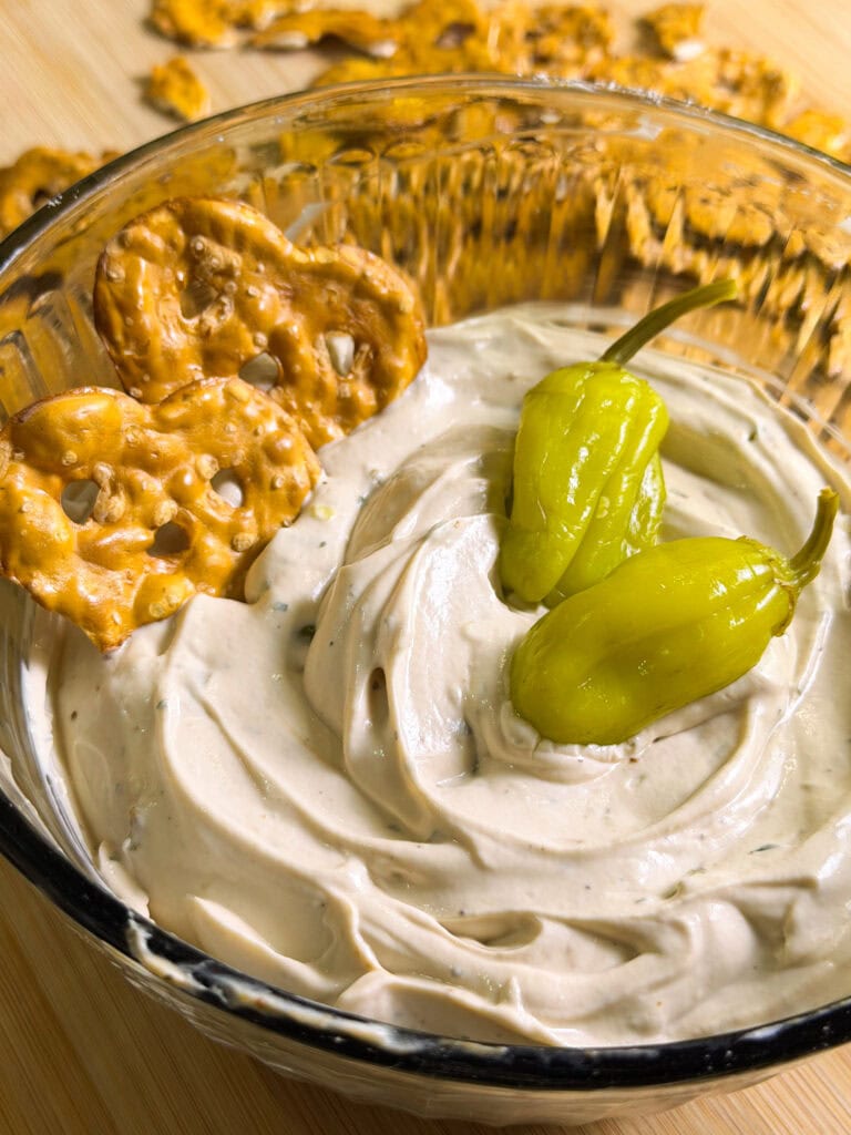 A glass bowl filled with creamy dip, topped with two whole pepperoncini peppers and two pretzel crisps. More pretzel crisps are scattered in the background on a wooden surface.