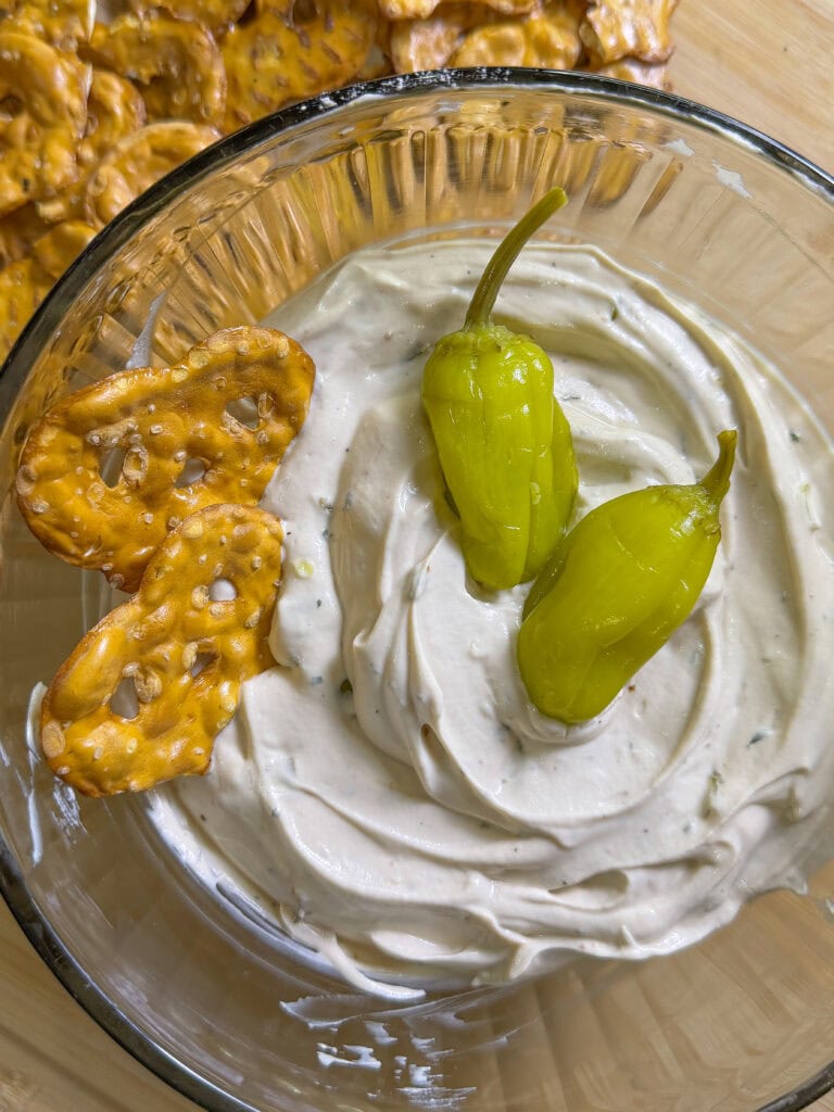 A glass bowl of creamy dip topped with two yellow-green pepperoncini peppers, with pretzel crisps partially dipped into the dip. More pretzel crisps are visible in the background.