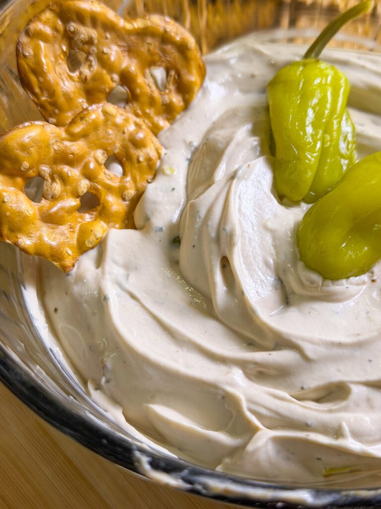 A close-up of a glass bowl filled with creamy dip, garnished with two yellow-green peppers and served with two pretzel crisps on the side. The dip has a smooth, swirled texture.