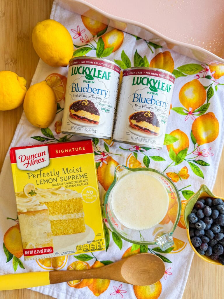 A box of lemon cake mix, two cans of blueberry filling, fresh lemons, a bowl of blueberries, a glass measuring cup with batter, a wooden spoon, and a colorful lemon-patterned towel are arranged on a wooden surface.