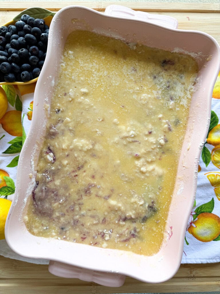 A baking dish with an unbaked blueberry cobbler mixture sits on a lemon-patterned cloth. Fresh blueberries and lemons are nearby on a wooden surface.
