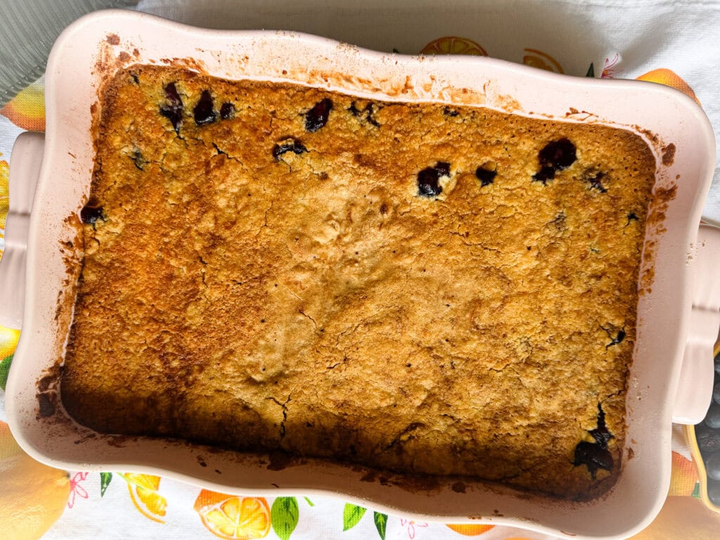 A golden-brown baked dessert with a slightly cracked top and visible dark berries, in a rectangular ceramic baking dish placed on a colorful, fruit-themed cloth.