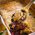 A close-up of a golden-brown berry cobbler in a baking dish, with a wooden spoon lifting a serving, showing gooey berries and syrupy filling. The dish sits on a patterned tablecloth with fruit designs.