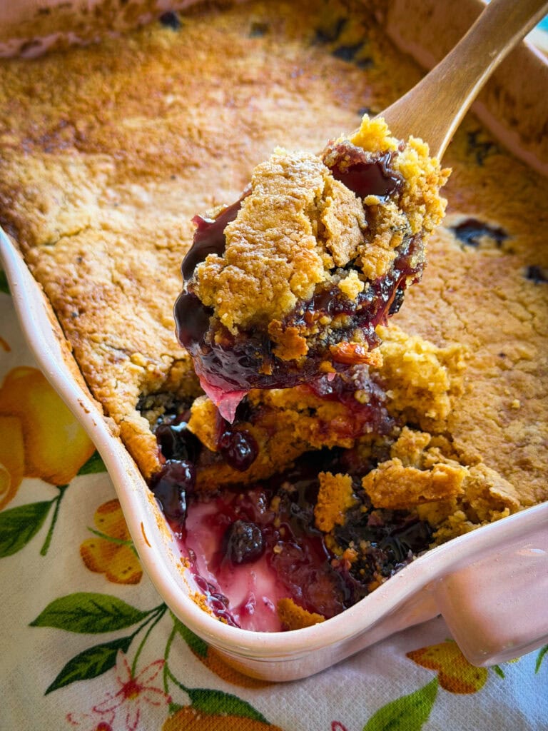 A close-up of a golden-brown berry cobbler in a baking dish, with a wooden spoon lifting a serving, showing gooey berries and syrupy filling. The dish sits on a patterned tablecloth with fruit designs.