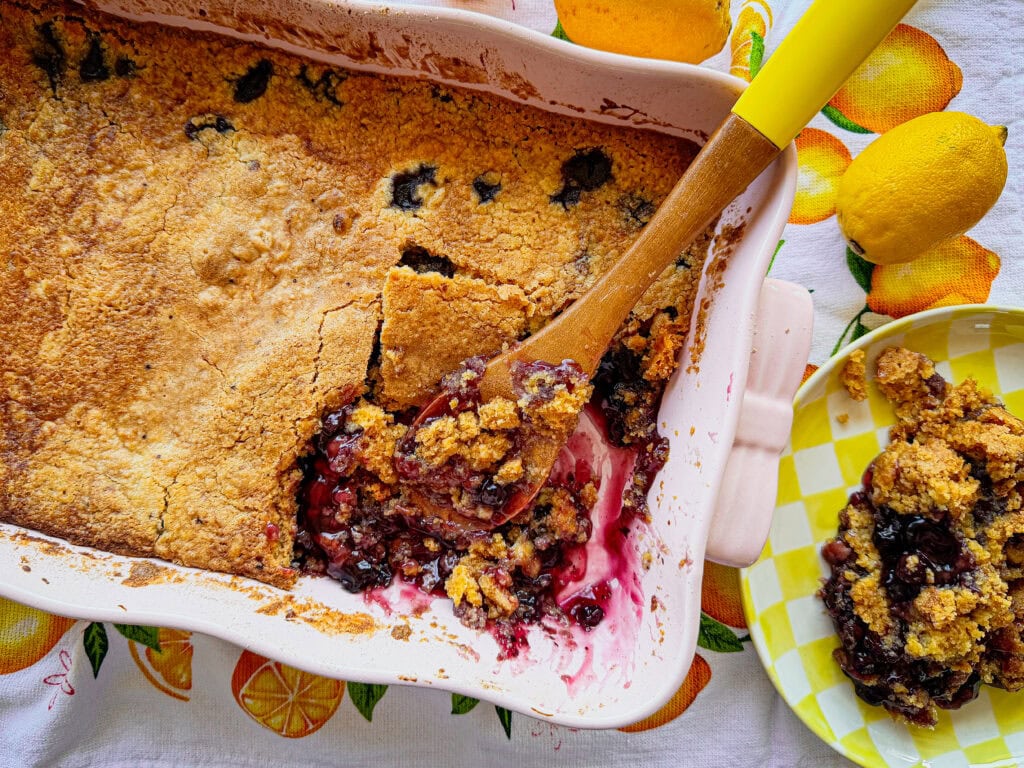 A freshly baked berry cobbler in a baking dish, with a wooden spoon serving a portion onto a yellow checkered plate. Lemons and a citrus-patterned tablecloth are in the background.