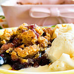 A close-up of a yellow plate with a serving of fruit cobbler and two scoops of vanilla ice cream, with a pink baking dish in the background. The dessert appears warm and crumbly with a golden topping.
