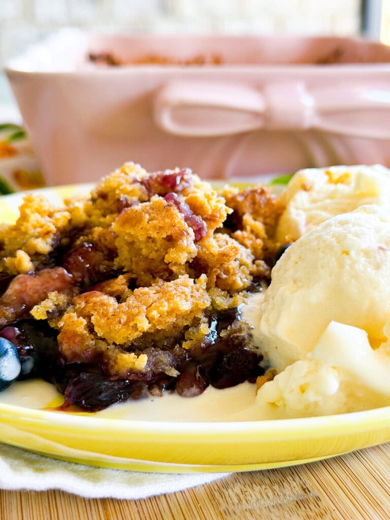 A close-up of a yellow plate with a serving of fruit cobbler and two scoops of vanilla ice cream, with a pink baking dish in the background. The dessert appears warm and crumbly with a golden topping.