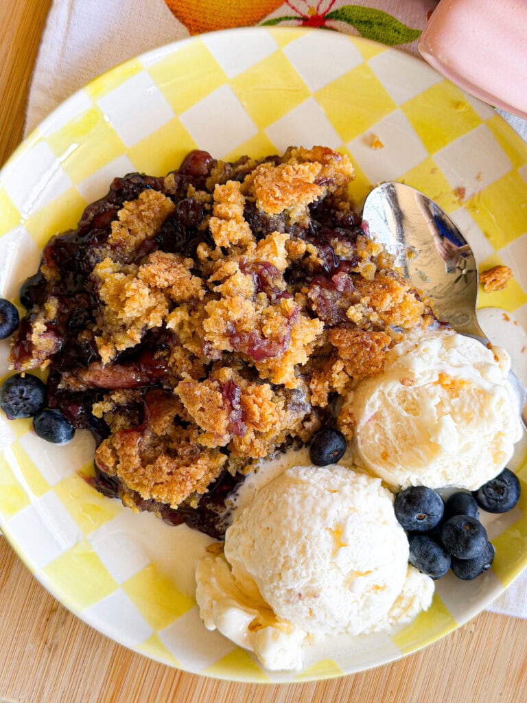 A plate with blueberry crumble, two scoops of vanilla ice cream, and fresh blueberries, served on a yellow-and-white checkered dish with a spoon beside the dessert.