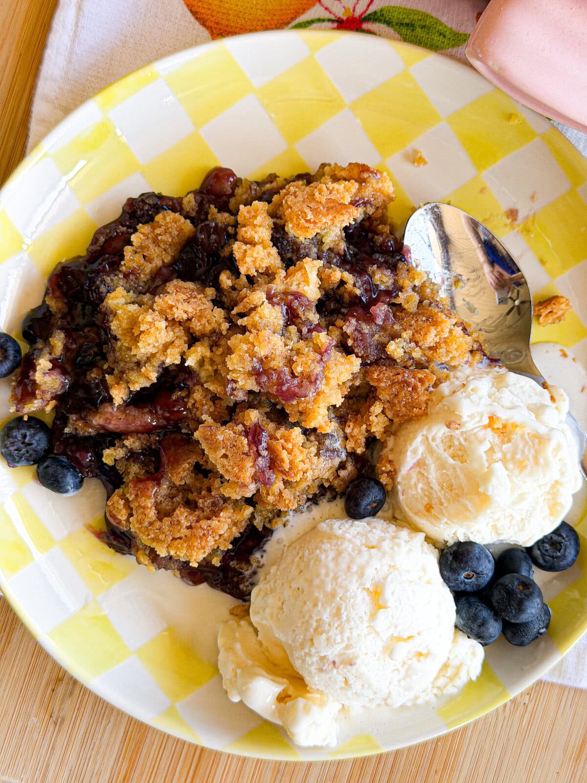 A plate with blueberry crumble, two scoops of vanilla ice cream, and fresh blueberries, served on a yellow-and-white checkered dish with a spoon beside the dessert.