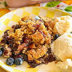 A plate of blueberry cobbler topped with golden crumble, served with two scoops of vanilla ice cream and fresh blueberries on a yellow checkered plate.