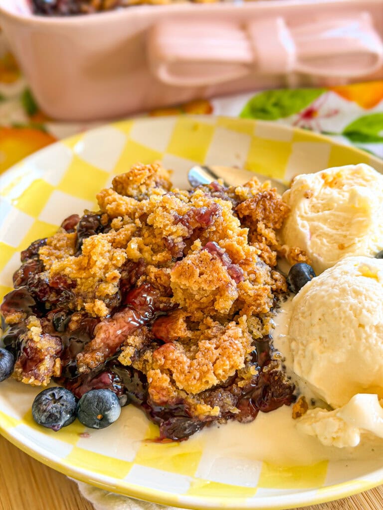 A plate of blueberry cobbler topped with golden crumble, served with two scoops of vanilla ice cream and fresh blueberries on a yellow checkered plate.