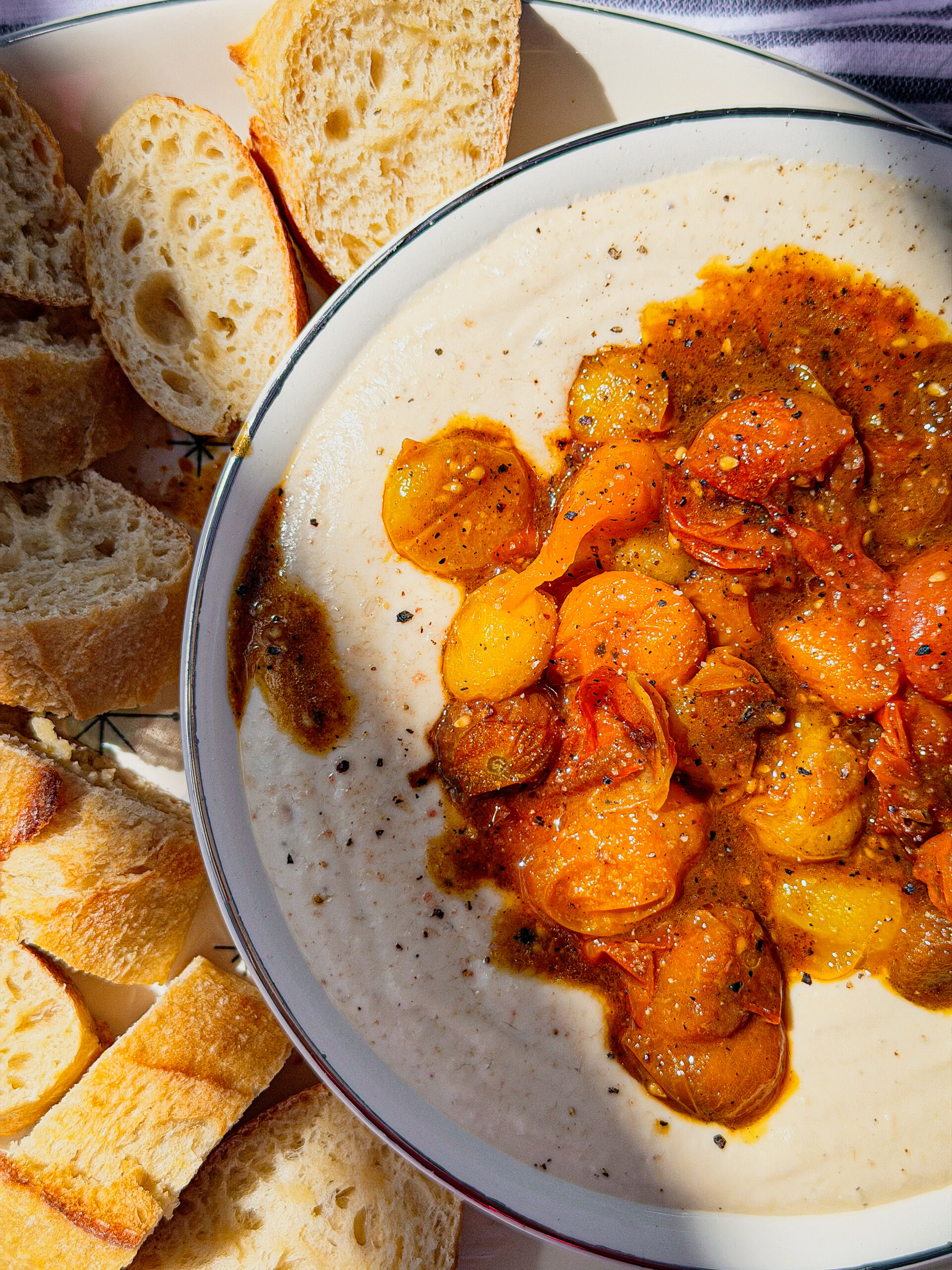 A bowl of creamy hummus topped with roasted yellow and orange cherry tomatoes and spices, surrounded by sliced crusty baguette bread. Bright sunlight highlights the food.