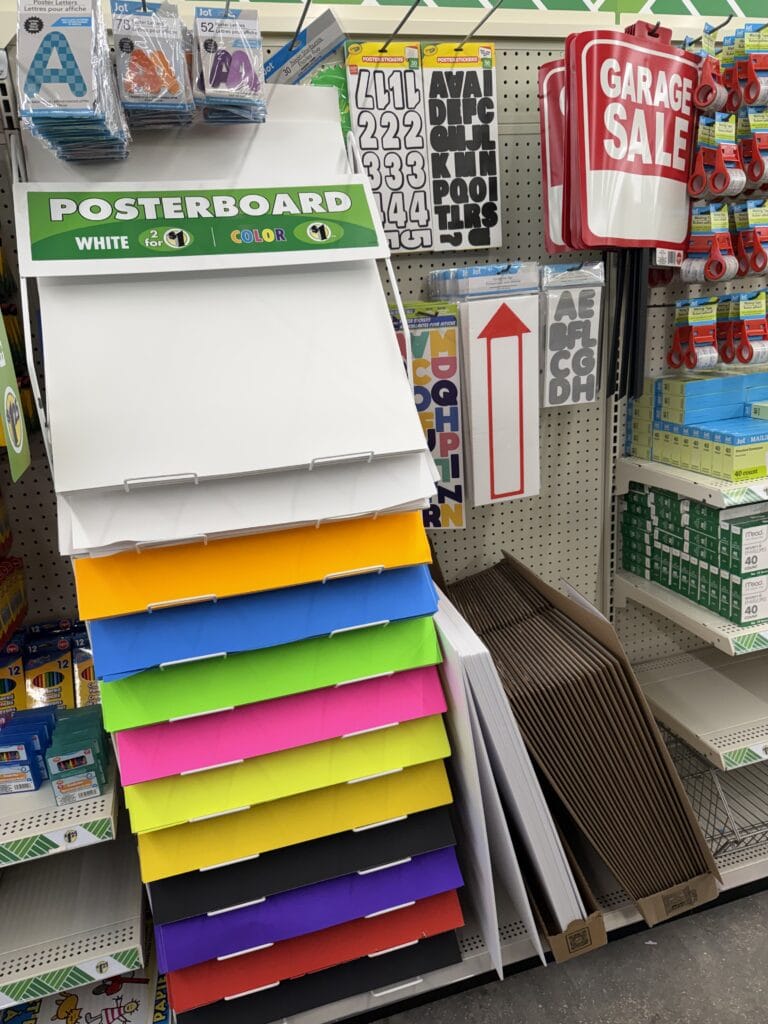 A store display with colorful poster boards stacked vertically, alphabet and number stickers, red Garage Sale signs, and arrow signs on shelves, along with other stationery items.