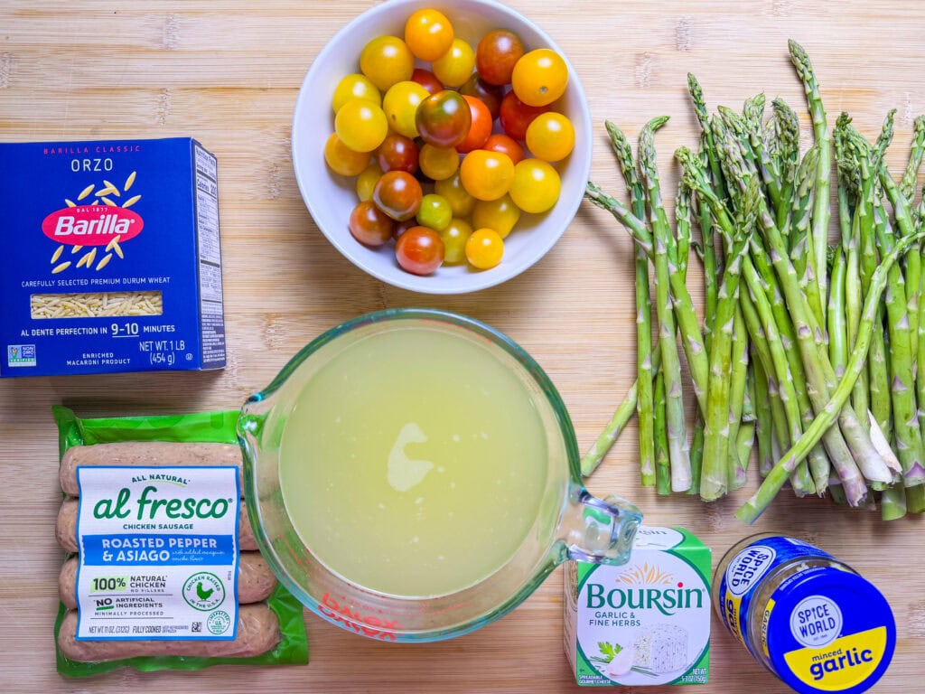 A top-down view of ingredients on a wooden surface: a box of orzo pasta, a bowl of cherry tomatoes, a bunch of asparagus, a pack of sausage, a measuring cup of broth, Boursin cheese, and a container of garlic powder.