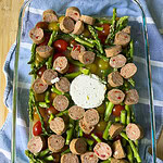 A glass baking dish with halved cherry tomatoes, chopped asparagus, sliced sausage, and a round of goat cheese in the center, resting on a blue and white striped cloth on a wooden surface.