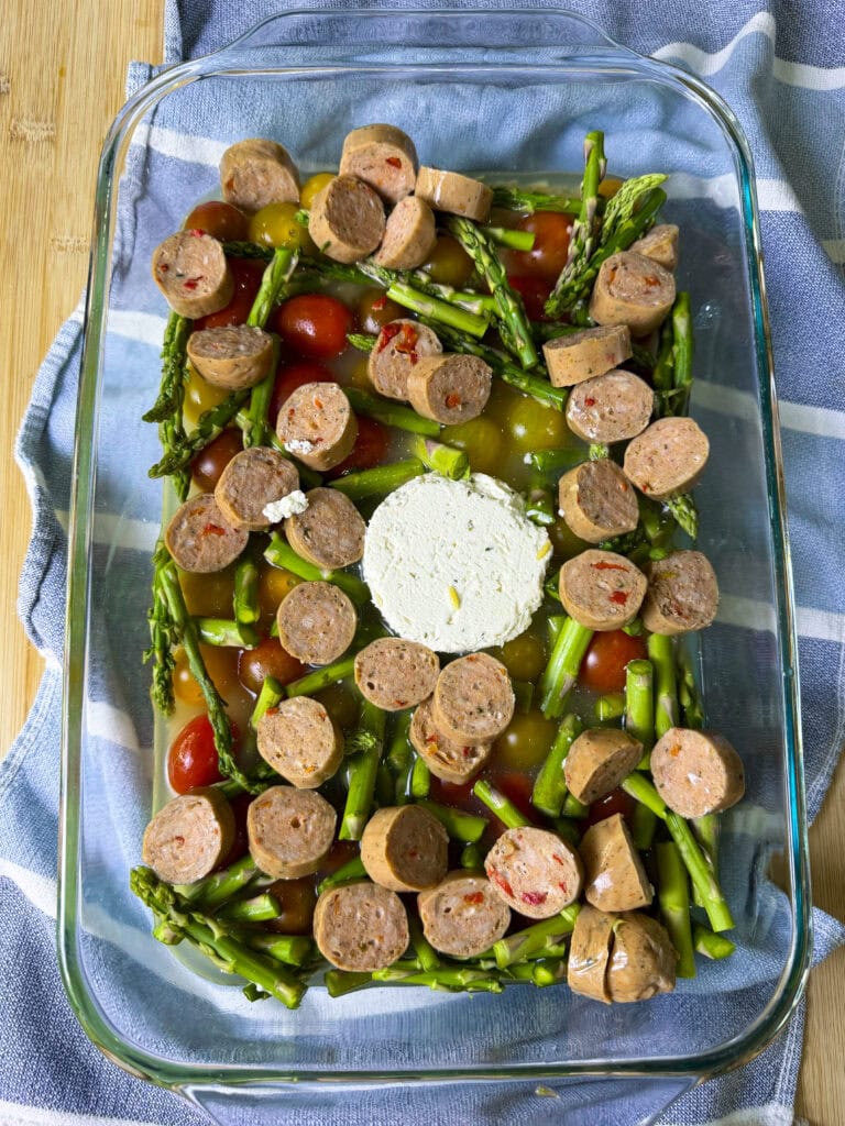 A glass baking dish with halved cherry tomatoes, chopped asparagus, sliced sausage, and a round of goat cheese in the center, resting on a blue and white striped cloth on a wooden surface.