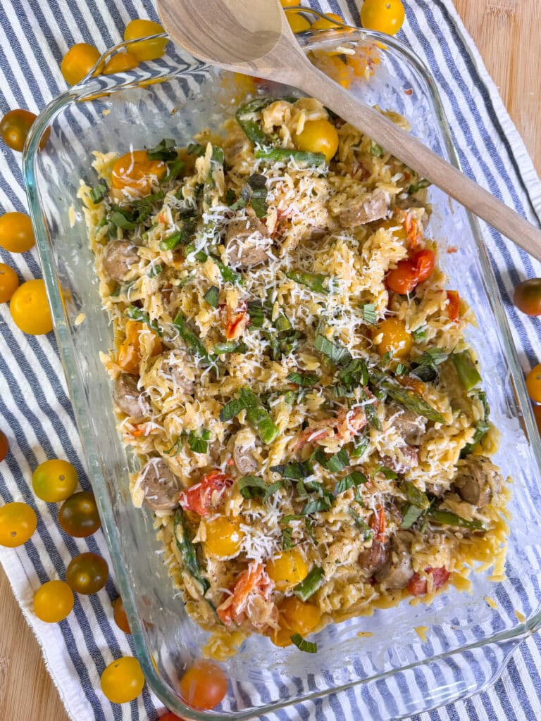 A glass baking dish filled with a baked pasta dish containing orzo, cherry tomatoes, green beans, shredded cheese, and herbs, sits on a striped cloth. A wooden spoon rests on the edge. Yellow and multicolored tomatoes are scattered nearby.