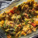A glass baking dish filled with baked orzo pasta, cherry tomatoes, sausage, asparagus, and topped with shredded cheese and fresh herbs, sits on a striped cloth in bright sunlight.